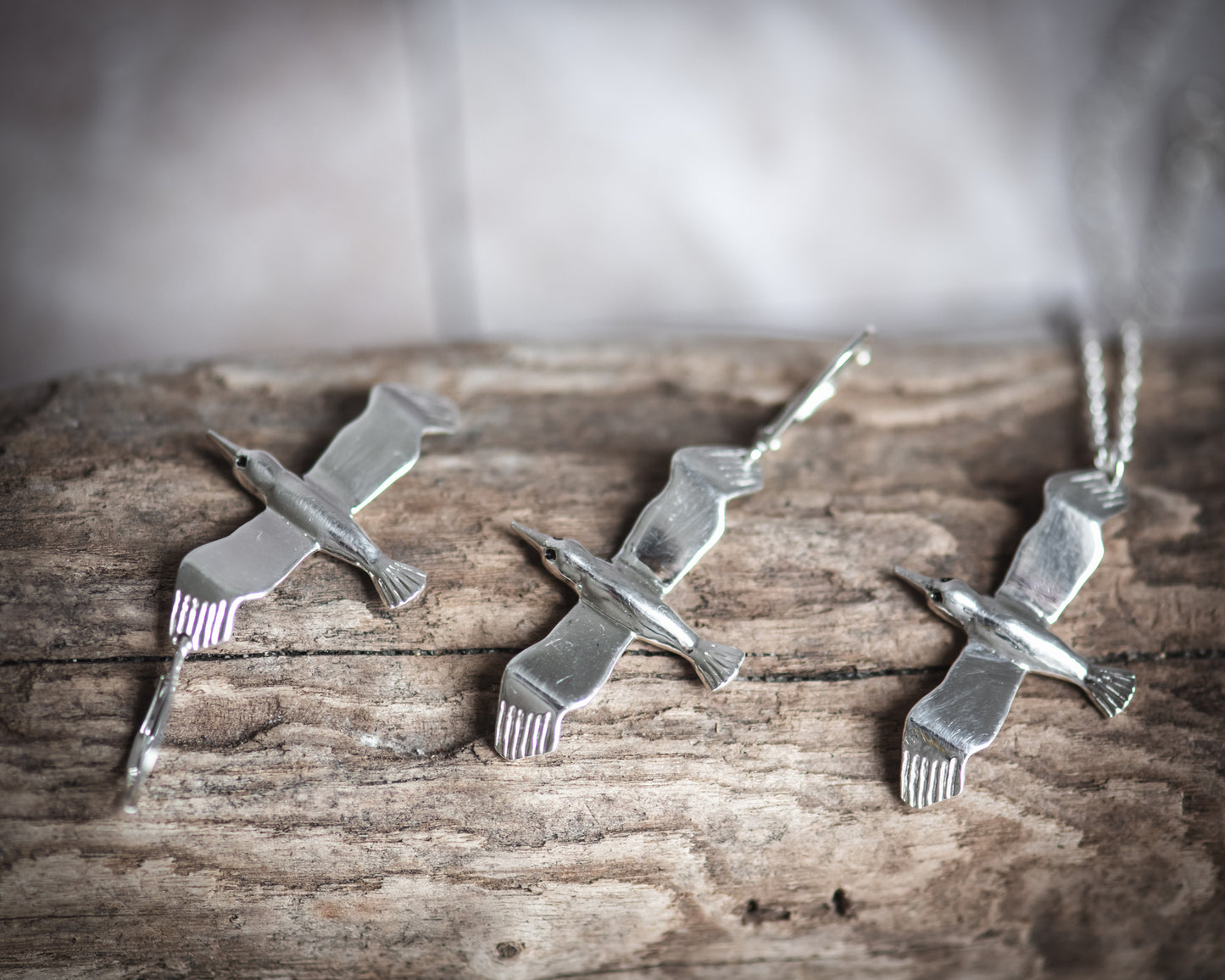 Three silver bird-shaped pendant earrings and an albatross necklace pendant on a wooden surface with a blurred background