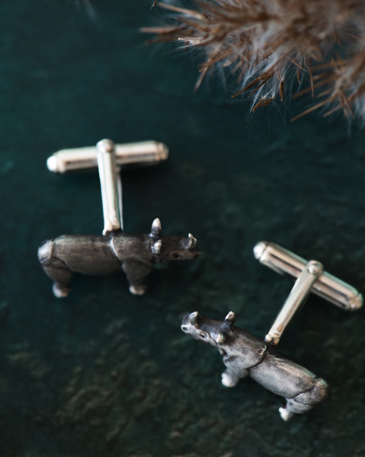 Silver rhinoceros cufflinks on a dark green surface with dried grasses.