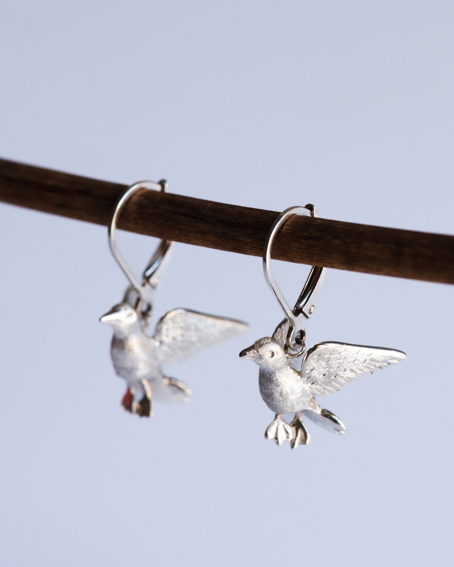Silver songbird earrings hanging from a branch against a light blue background