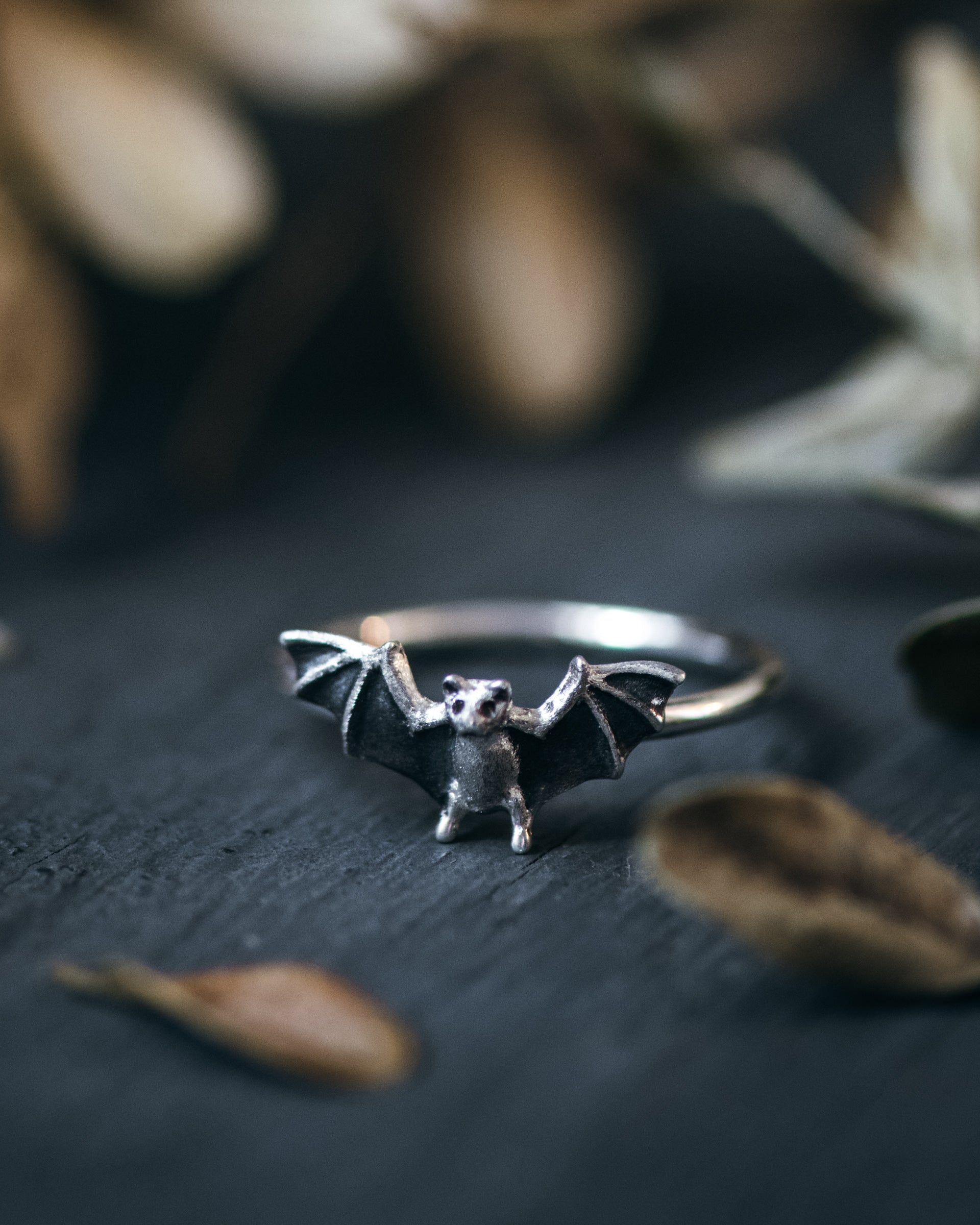 Silver bat-shaped ring on a dark surface with dried leaves
