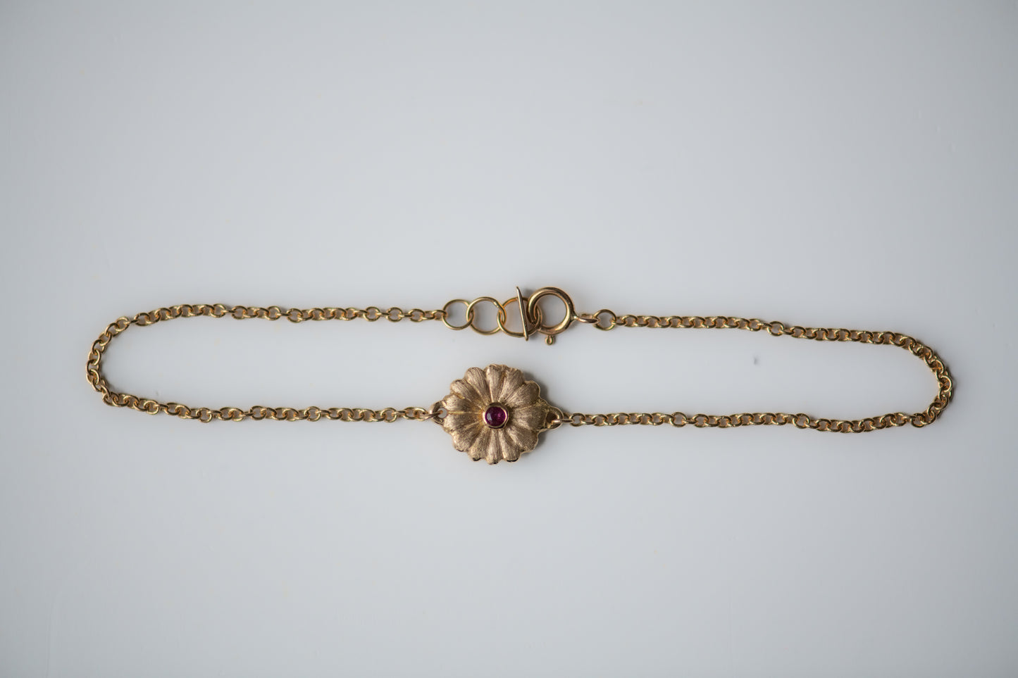 Gold bracelet with a daisy pendant and a ruby on a light gray background