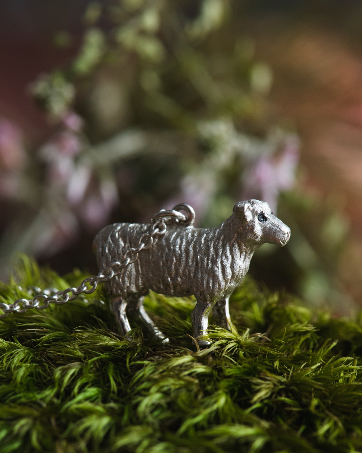 Silver sheep-shaped pendant on a chain placed on green moss with a blurred natural background.