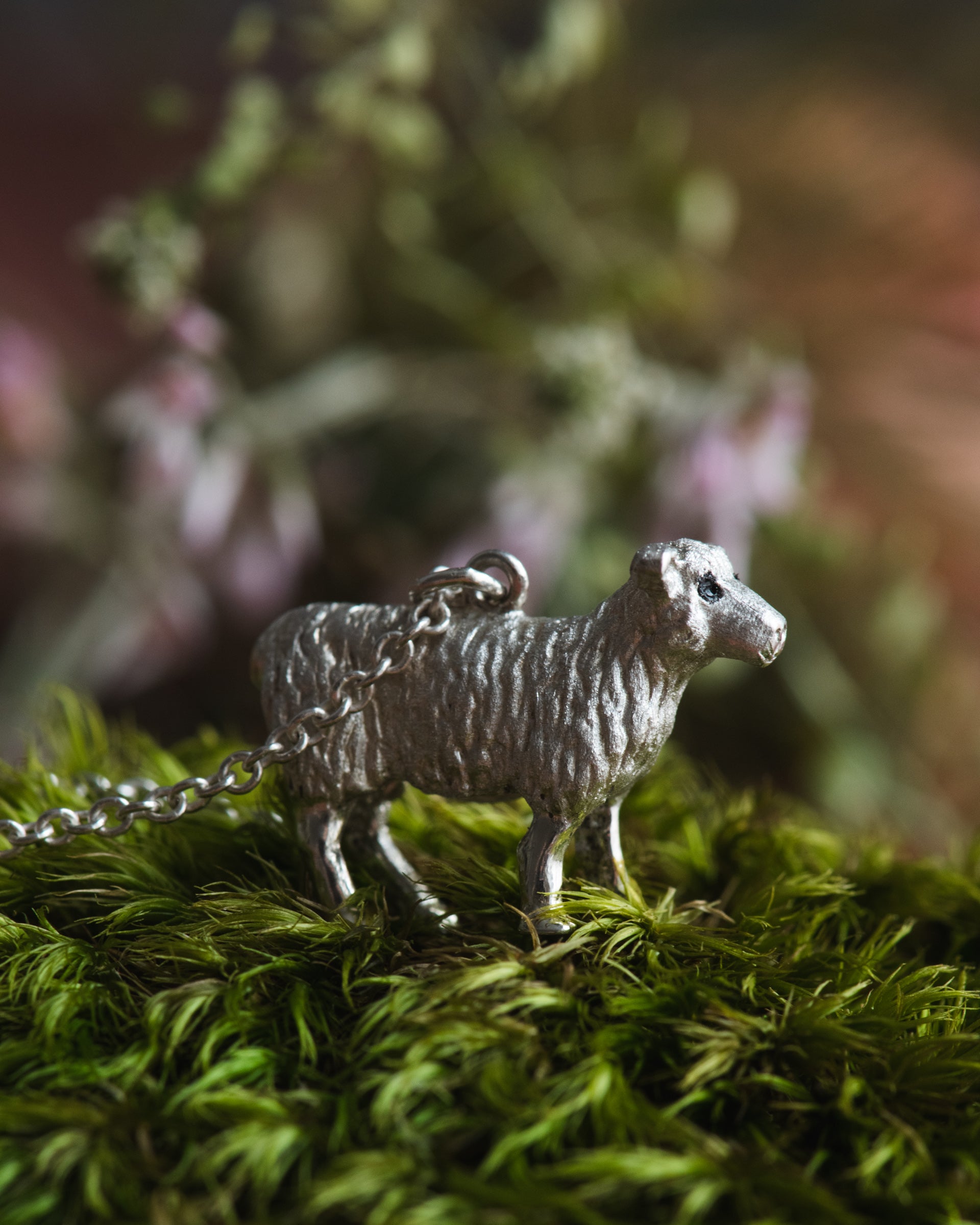 Silver sheep-shaped pendant on a chain placed on green moss with a blurred natural background.