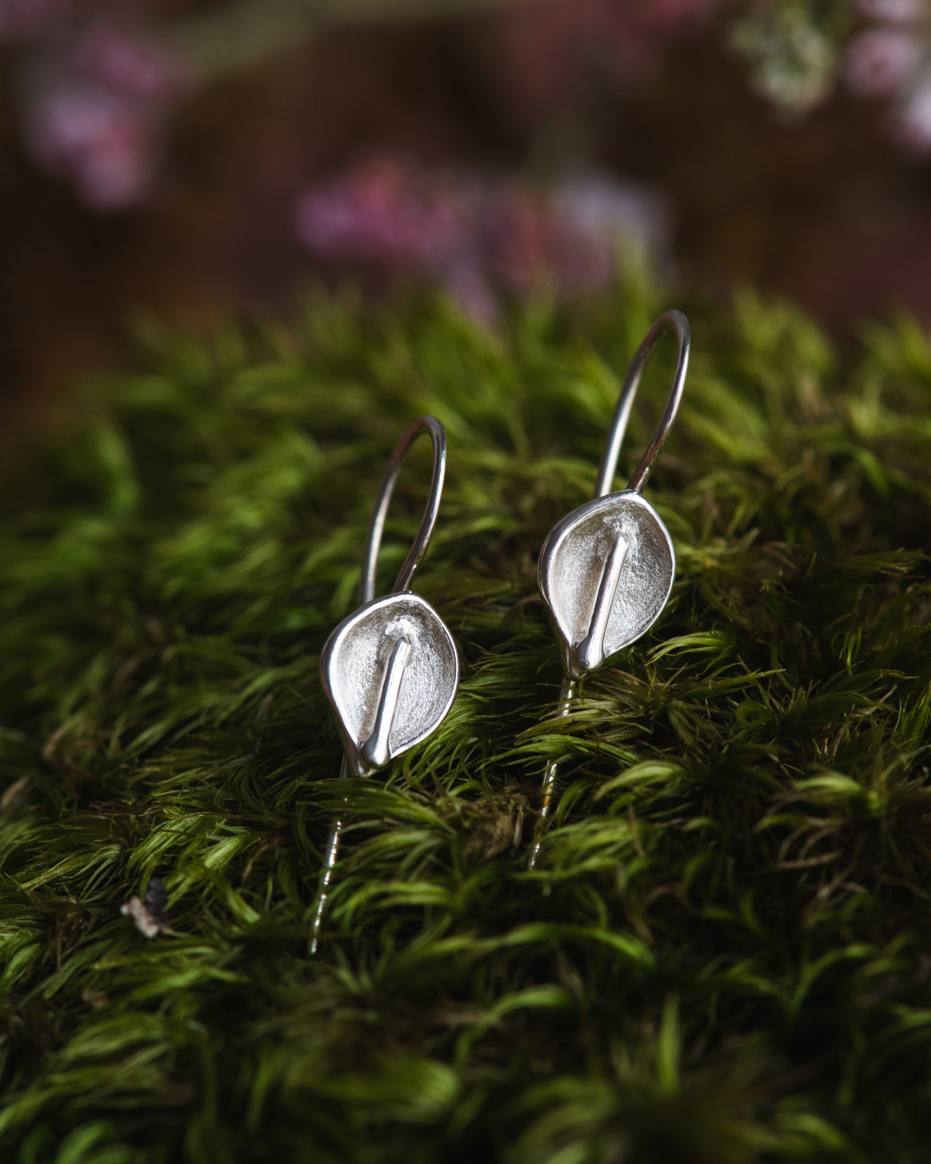Silver calla lily earrings on a bed of green moss with blurred pink flowers in the background.