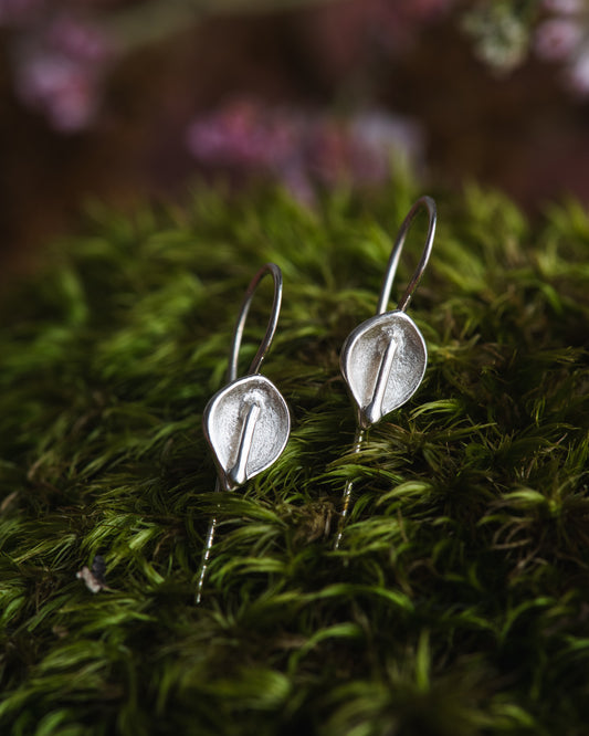 Silver calla lily earrings on a bed of green moss with blurred pink flowers in the background.
